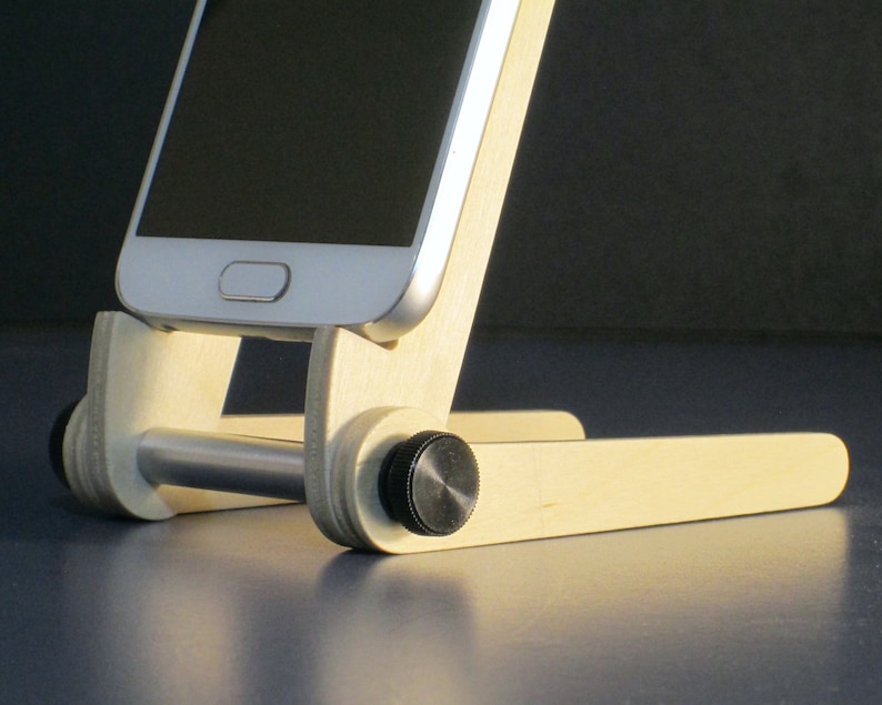 May include: A wooden phone stand with a silver metal bar and black knobs. A white smartphone is placed on the stand. The stand is made of light-colored wood and has a simple, functional design.