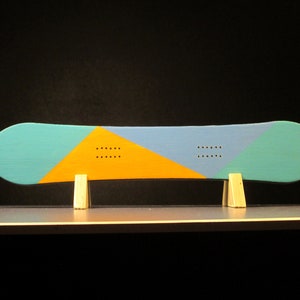 May include: A snowboard with a teal, blue, and yellow geometric design. The board is supported by two small wooden stands. The design includes a series of small, evenly spaced holes. The background is black.