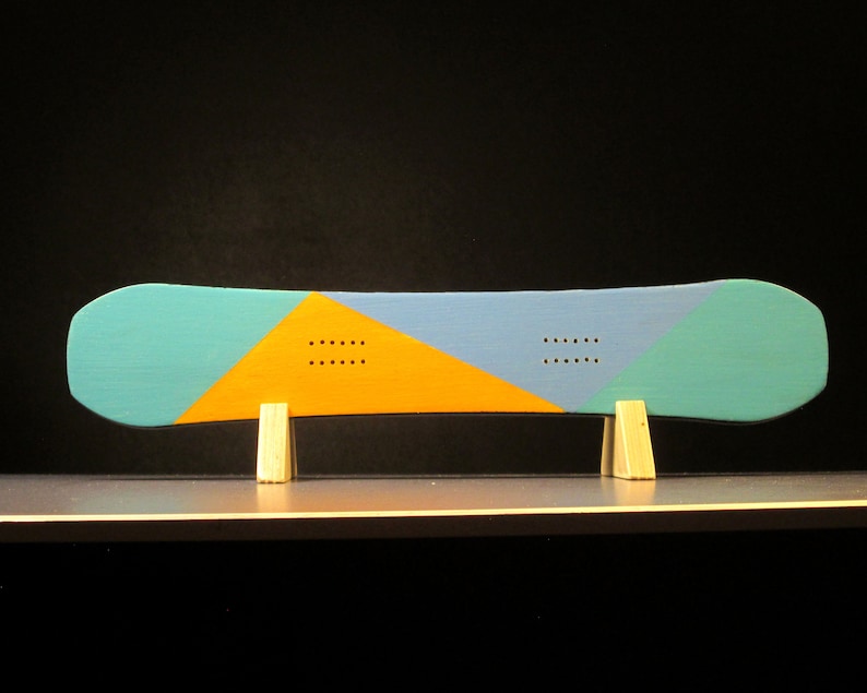 May include: A snowboard with a teal, blue, and yellow geometric design. The board is supported by two small wooden stands. The design includes a series of small, evenly spaced holes. The background is black.