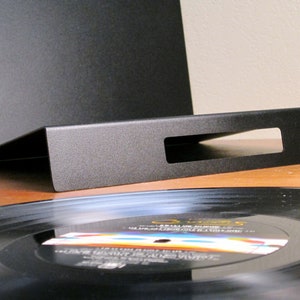 May include: A black metal record holder with a rectangular cutout, angled to display vinyl records. A black vinyl record is partially visible, resting on a wooden surface. The holder is designed to showcase and store vinyl records.