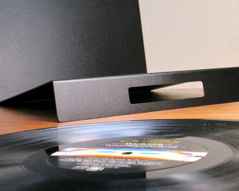 May include: A black metal record holder with a rectangular cutout, angled to display vinyl records. A black vinyl record is partially visible, resting on a wooden surface. The holder is designed to showcase and store vinyl records.