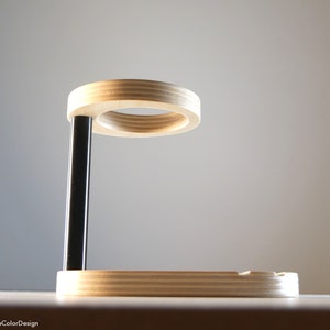 May include: A modern wooden phone stand with a circular top and a rectangular base. The stand has a black support beam and is made of light-colored wood. The design is minimalist and functional, suitable for a desk or bedside table.