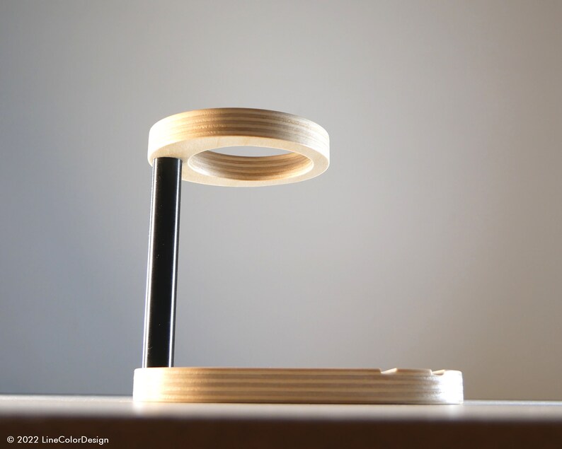 May include: A modern wooden phone stand with a circular top and a rectangular base. The stand has a black support beam and is made of light-colored wood. The design is minimalist and functional, suitable for a desk or bedside table.