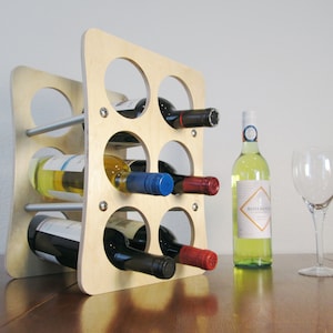 May include: A light-colored wooden wine rack with six circular openings, holding various wine bottles. A bottle of white wine and a wine glass are next to the rack. The rack is made of light-colored wood and has metal rods.