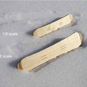 May include: Two miniature wooden snowboards, one labeled 1/6 scale and the other 1/8 scale, rest in the snow. The snowboards are light brown with rounded tips and a series of small holes. The text "1/8 scale" and "1/6 scale" are visible.