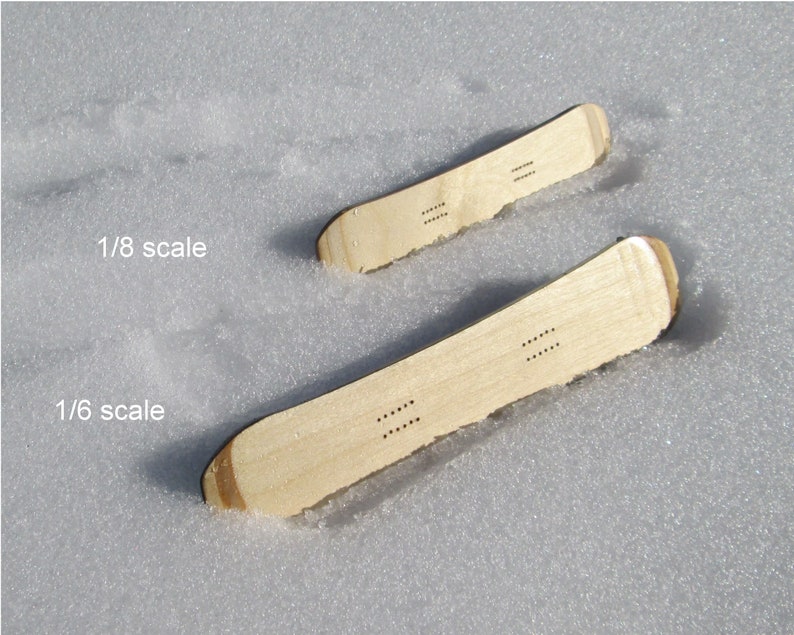 May include: Two miniature wooden snowboards, one labeled 1/6 scale and the other 1/8 scale, rest in the snow. The snowboards are light brown with rounded tips and a series of small holes. The text "1/8 scale" and "1/6 scale" are visible.
