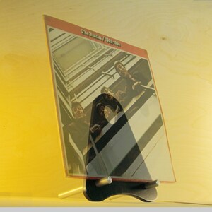 May include: A vinyl record album titled "The Beatles / 1962-1966" displayed in a black and silver stand. The album cover features a photograph of the band members. The stand is on a light-colored wooden surface.