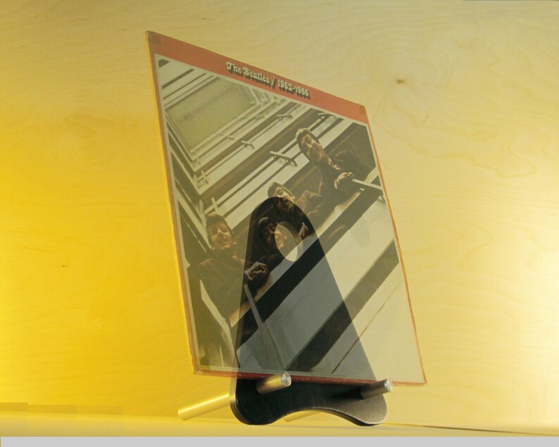 May include: A vinyl record album titled "The Beatles / 1962-1966" displayed in a black and silver stand. The album cover features a photograph of the band members. The stand is on a light-colored wooden surface.