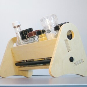 May include: A light-colored wooden spice rack with a unique angled design. It holds various spice jars and bottles, including a clear pepper grinder. The rack has a handle and decorative cutouts, and a black knob.