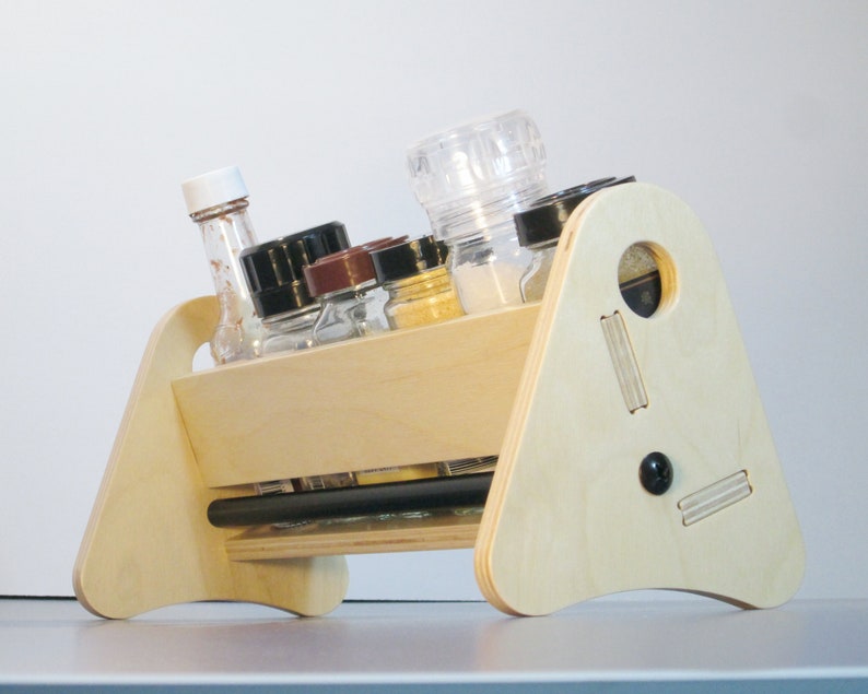 May include: A light-colored wooden spice rack with a unique angled design. It holds various spice jars and bottles, including a clear pepper grinder. The rack has a handle and decorative cutouts, and a black knob.
