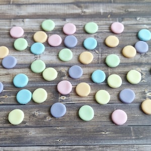 May include: A collection of pastel-colored macarons in shades of pink, yellow, green, blue, and purple are scattered on a dark wooden surface. The round, flat cookies are arranged in a random pattern, creating a visually appealing display.