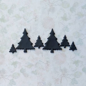 May include: A collection of seven black, stylized Christmas tree shapes arranged in a row. The trees vary in size, with the largest in the center. The background is a light, patterned surface with floral and leaf designs.