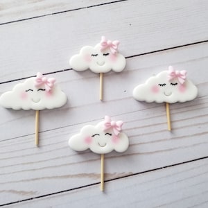 May include: Four white cloud-shaped decorations with pink bows and smiling faces on wooden sticks. The clouds have rosy cheeks and closed eyes. These are likely cake toppers or party decorations.
