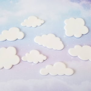 May include: Seven white cloud-shaped cake decorations on a light purple background with a sparkly texture.