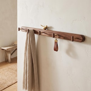May include: A long, horizontal wall-mounted coat rack made of dark brown wood with rounded edges. It features five small, cylindrical pegs and a decorative brass bird figurine perched on top. A beige scarf and a leather keychain hang from the pegs.