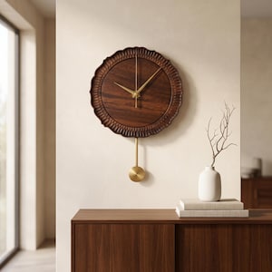 May include: A round wall clock featuring a dark brown wood grain face with a scalloped, carved wooden frame. The clock has thin, metallic gold hands and a matching gold pendulum that hangs below the circular body, creating a balanced design.