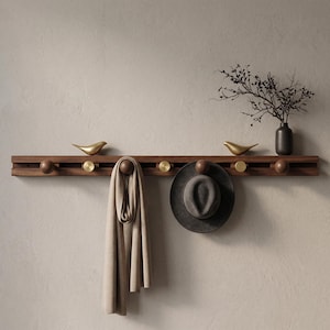 May include: A horizontal dark wood wall-mounted coat rack featuring alternating round wooden pegs and flat circular brass accents. Two gold-toned bird figurines sit on the top edge, and a beige scarf and a dark gray felt hat hang from the pegs.