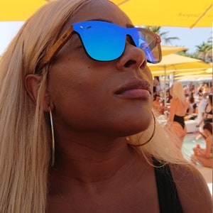 Puede incluir: Una mujer con el pelo largo y rubio que lleva gafas de sol azules espejadas con montura de madera. Lleva un top negro de tirantes y un aro de plata en la oreja izquierda.