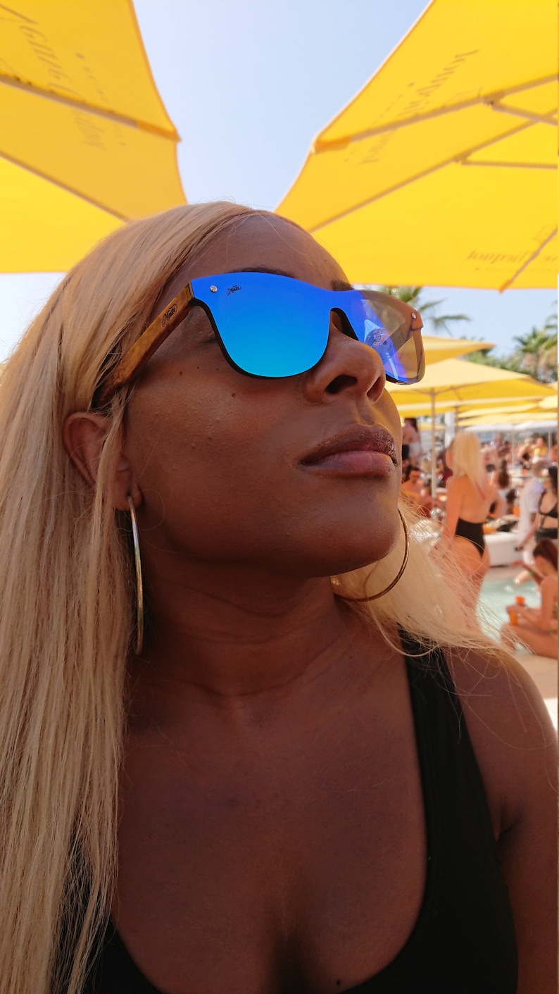 Puede incluir: Una mujer con el pelo largo y rubio que lleva gafas de sol azules espejadas con montura de madera. Lleva un top negro de tirantes y un aro de plata en la oreja izquierda.