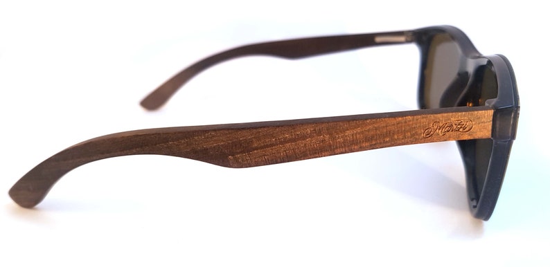Puede incluir: Vista lateral de unas gafas de sol de madera marr&oacute;n con monturas negras y lentes oscuras. Las gafas de sol tienen la palabra "Marley" grabada en el lateral de la montura.