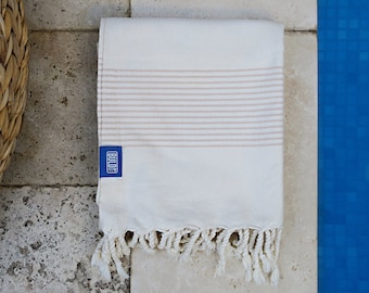 May include: Folded white towel with beige horizontal stripes and fringe. A blue label with white text is attached to the towel. The towel is placed on a light-colored stone surface next to a pool.