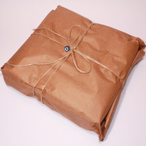 May include: A square package wrapped in brown paper, tied with twine, and adorned with a blue and white evil eye charm. The package is neatly folded and presented on a white surface.