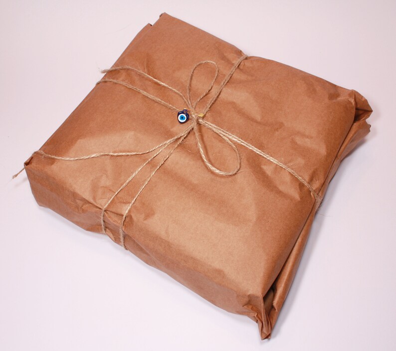 May include: A square package wrapped in brown paper, tied with twine, and adorned with a blue and white evil eye charm. The package is neatly folded and presented on a white surface.