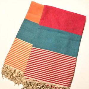 May include: A colorful rectangular beach towel with a patchwork design. The towel features sections of orange, teal, and red. The bottom half has striped patterns in orange and red. The towel has a fringed edge.