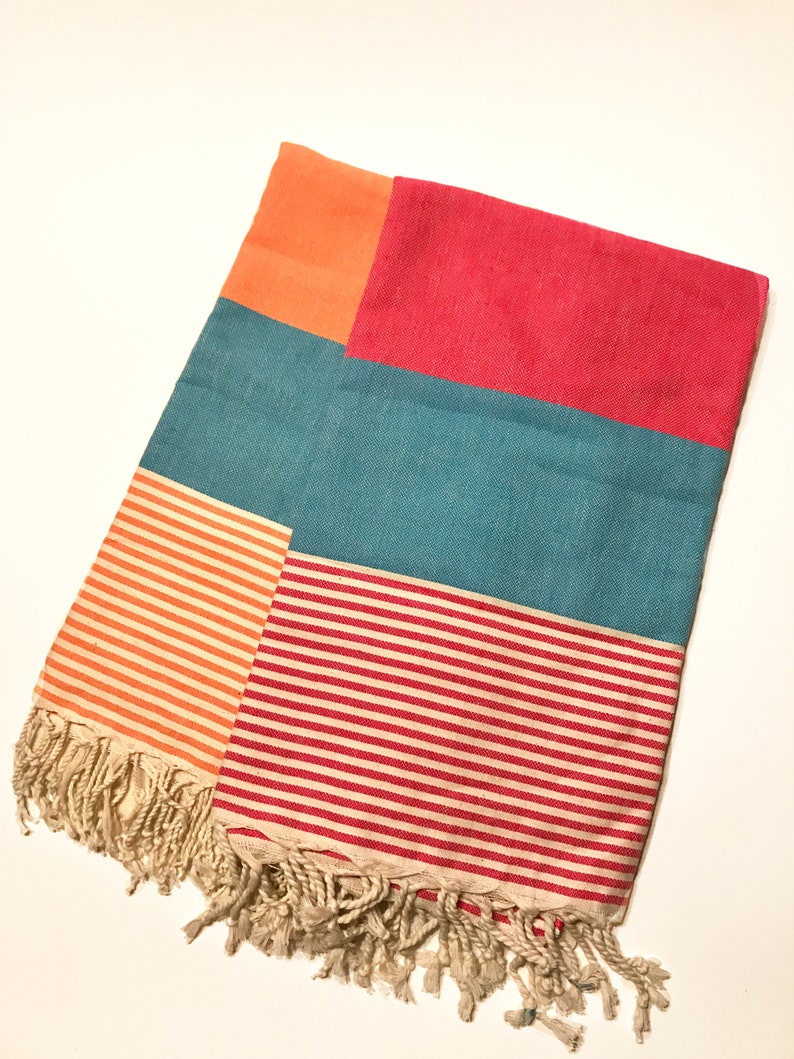 May include: A colorful rectangular beach towel with a patchwork design. The towel features sections of orange, teal, and red. The bottom half has striped patterns in orange and red. The towel has a fringed edge.