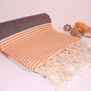May include: Rolled beach towel with orange and white stripes, a dark gray border, and cream-colored fringe. The towel is partially unrolled, revealing the striped pattern. Several seashells are arranged nearby.
