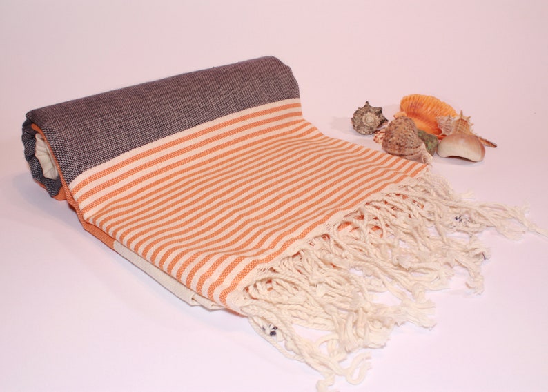 May include: Rolled beach towel with orange and white stripes, a dark gray border, and cream-colored fringe. The towel is partially unrolled, revealing the striped pattern. Several seashells are arranged nearby.