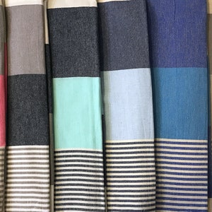 May include: A collection of folded, colorful striped beach towels. Each towel features a solid color block at the top, a contrasting stripe in the middle, and a striped pattern at the bottom. Colors include orange, pink, gray, teal, blue, and more.