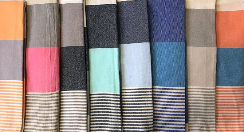 May include: A collection of folded, colorful striped beach towels. Each towel features a solid color block at the top, a contrasting stripe in the middle, and a striped pattern at the bottom. Colors include orange, pink, gray, teal, blue, and more.