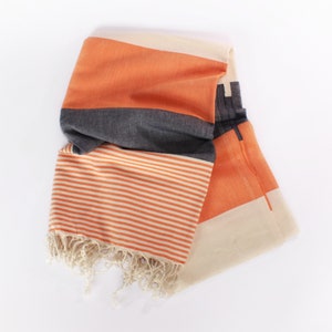 May include: Folded orange, gray, and cream-colored striped towel with fringe. The towel features a geometric design with orange and cream-colored stripes and a gray band. The fringe is a natural cream color.