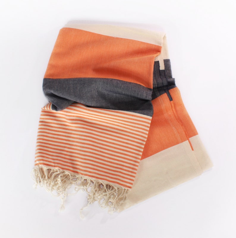 May include: Folded orange, gray, and cream-colored striped towel with fringe. The towel features a geometric design with orange and cream-colored stripes and a gray band. The fringe is a natural cream color.