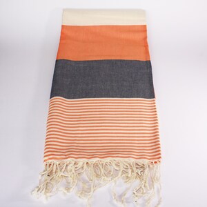 May include: A rectangular beach towel with a cream-colored top, followed by orange, dark gray, and orange and white striped sections. The bottom edge has cream-colored fringe. The towel is made of a soft, woven material.