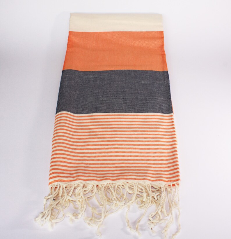 May include: A rectangular beach towel with a cream-colored top, followed by orange, dark gray, and orange and white striped sections. The bottom edge has cream-colored fringe. The towel is made of a soft, woven material.