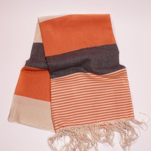 May include: A folded orange and cream-colored scarf with a striped pattern and fringe. The scarf has a solid orange section, a dark gray section, and a cream-colored section. The striped section features orange and cream-colored horizontal stripes.