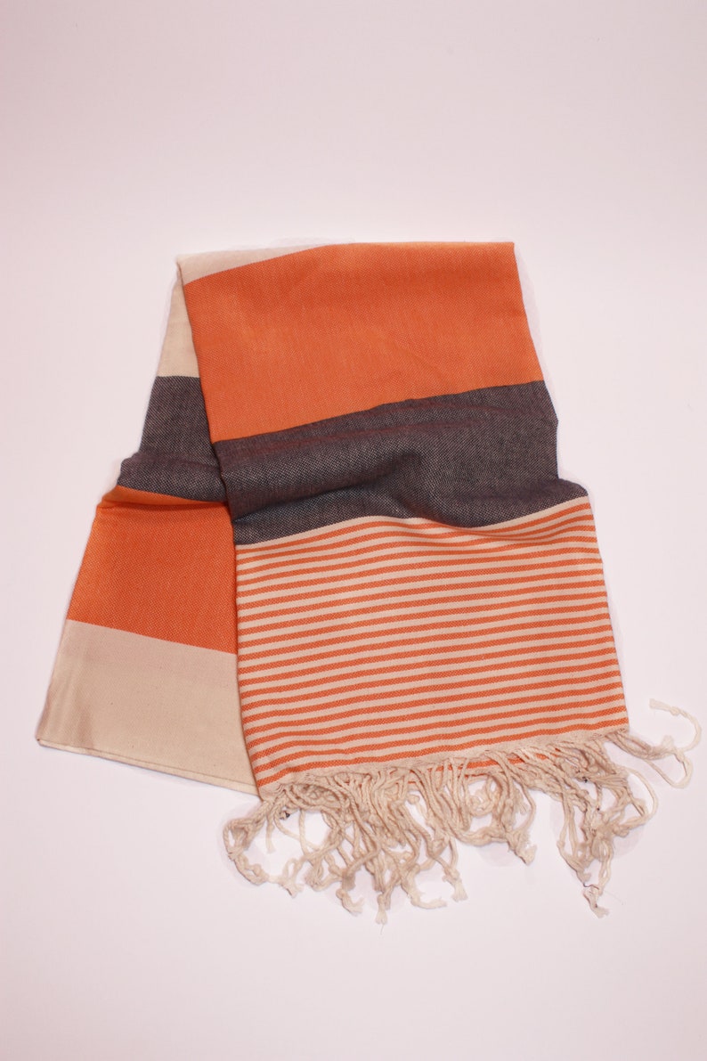 May include: A folded orange and cream-colored scarf with a striped pattern and fringe. The scarf has a solid orange section, a dark gray section, and a cream-colored section. The striped section features orange and cream-colored horizontal stripes.