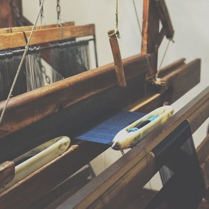 May include: A vintage wooden weaving loom with dark blue fabric in progress. The loom features a shuttle and various wooden components, showcasing the craftsmanship of textile production. The image has a warm, rustic aesthetic.