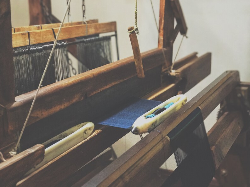 May include: A vintage wooden weaving loom with dark blue fabric in progress. The loom features a shuttle and various wooden components, showcasing the craftsmanship of textile production. The image has a warm, rustic aesthetic.