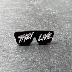 以下が含まれることがあります： 白い文字で「They Live」と書かれた、サングラスの形をした黒いエナメルピン