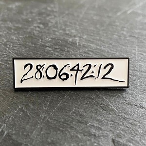 Puede incluir: Un pin de esmalte blanco con borde negro. El pin presenta el texto "28:06:42:12" en un estilo de escritura a mano.