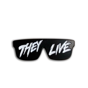 Può includere: Spilla in smalto nero a forma di occhiali da sole con il testo "THEY LIVE" in lettere bianche.