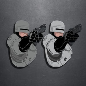 Può includere: Due adesivi identici con un'illustrazione stilizzata di un personaggio robotico che indossa un casco e un'armatura argentati, che tiene una pistola nera puntata in avanti. Il personaggio ha un mento e una bocca umani visibili.