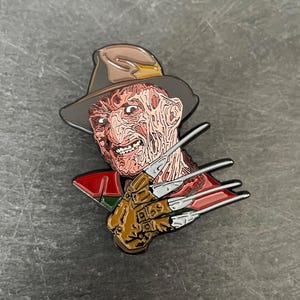 Pode incluir: Um broche de esmalte preto e vermelho com Freddy Krueger da franquia de filmes A Hora do Pesadelo. Ele está usando um chapéu marrom e tem uma expressão ameaçadora. Ele está segurando uma luva com lâminas afiadas como navalhas.