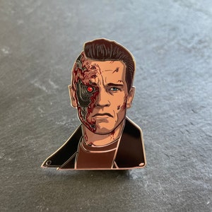 Może przedstawiać: Złota metalowa przypinka z szczegółową ilustracją cyborga z czerwonym okiem i częściowo odsłoniętą robo-twarzą. Cyborg nosi czarną skórzaną kurtkę.