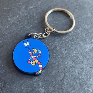 Puede incluir: Un llavero circular azul con una ilustración de dibujos animados de una hebra de ADN. El llavero tiene un anillo y una cadena de metal plateado. La hebra de ADN está compuesta por formas coloridas, incluyendo rojo, amarillo y blanco.