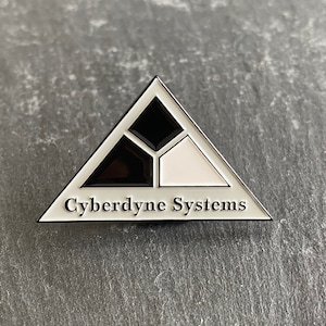 Könnte beinhalten: Ein weißer dreieckiger Pin mit einem schwarz-weißen geometrischen Design und dem Text "Cyberdyne Systems".