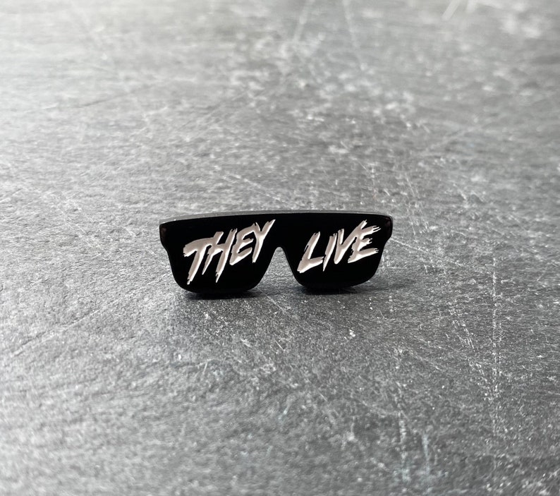 以下が含まれることがあります： 白い文字で「They Live」と書かれた、サングラスの形をした黒いエナメルピン